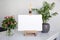 Blank canvas mockup on wooden easel and roses and houseplant on white table