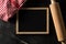 Blank, empty, black chalkboard with wooden rolling pin, wire whisk and red checkered dish towel flat lay from above on black