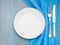 Blank flat white plate, fork, knife on blue wooden table, top view. Mock up, concept for marine restaurant, fish menu