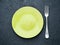 Blank flat yellow plate, fork on dark gray stone concrete table, top view. Mock up, copy space