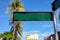 Blank street sign on sunny urban landscape with palm trees and yellow building. Green metallic sign plate on pillar