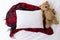 Blank white rectangle cushion laying on a tartan Christmas scarf with a white background and child`s teddy
