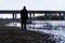 A bleak, moody, winter edit of a figure standing next to a lake, looking out to a motorway bridge, out of focus in the background
