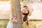 Blissful brown-eyed girl in trendy boater with white ribbon holding by tree and laughing. Outdoor portrait of ecstatic