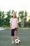 A blond 6 year old boy stands on a green lawn on a soccer field with a soccer ball