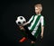 Blond boy child in sports green and white clothes playing soccer ball over dark background. Trendy sports