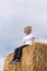 Blond boy in jeans and white T-shirt sits in the hay and looks into the distance. Portrait of child on haystack