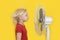 Blond boy stands near ventilator on bright yellow background. Hot summer