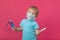 A blond child with an inhaler and a thermometer in his hand on a plain pink background