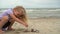 Blond girl playing with pebble stones on seashore. Lovely child with curly hair