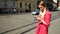 Blond girl in red coat standing on the street, typing on phone