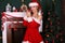 Blond girl wears Santa costume,posing beside Christmas tree and chimney