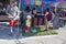 Blond haired child mowing a patch of artifical grass in front of a taco stand with a toy lawnmower while adults are buying food at