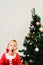 Blond and handsome 5 year old boy dressed as Santa Claus bored by the Christmas tree while waiting for the arrival of the real