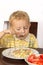 Blond little boy trying to eat with a fork potatoes with meat and tomatoes