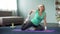 Blond middle-aged female doing stretching exercises on yoga mat at home, sport