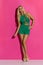 Blond Woman In Green Short Jumpsuit And High Heels Against Pink Wall
