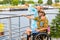 Blond woman standing with man in wheelchair by river and pointing at something