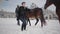 Blond woman and tall man leading two brown horses at the snow winter ranch. One stubborn animal stopped and wants to go