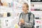 Blond young female traveler wearing travel backpack choosing perfume in airport duty free store.