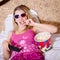 Blond young woman in color pajamas lying in bed with TV remote control in hand watching movie in 3D glasses and eating popcorn