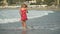 Blond young woman in short red dress spinning around, standing in shell seawater. Cute girl spends time at the sea