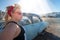 Blonde adult female with a 1950s vintage pin up hairstyle stands near an abandoned car