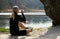 Blonde athlete girl sitting on ground to relax after jogging drinking water under a tree on a lake shore