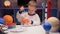 Blonde boy painting planet Mars of solar system sitting at table in evening with planets of solar system and space