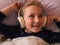 Blonde child girl in headphones learning language listening to music podcast with smartphone online in bed at home