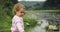 Blonde curly little girl throw stones in the river. Mountains background.