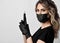 Blonde curly woman in black protective medical mask and latex gloves stands sideways holding permanent makeup machine