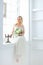 Blonde european bride sitting near window with bouquet of flowers.