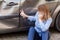 Blonde female in casual attire sitting near dented car and touching scratches and dents on automobile door, talking phone with