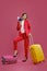 Blonde female in red pantsuit, white blouse, black heels. She put her leg on suitcase, yellow one nearby, holding