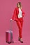 Blonde female in red pantsuit, white blouse, high black heels. She smiling, posing with suitcase on pink background