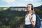 Blonde female traveler  with backpack stands on top of mountain on backdrop blue sky, active tourist woman with headphones hiking