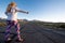 Blonde female wearing patriotic American clothing stands in the middle of a mountain road with her thumb out, concept for