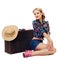 Blonde girl in checkered shirt and denim shorts is sitting near an old suitcase with straw hat and smiling playfully at