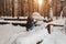 A blonde girl in a gray hat and a dark blue jacket in the winter in the forest is sitting in a snowdrift and throws her snow into