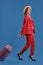 Blonde girl in hat, white blouse, red pantsuit, high heels. She carrying pink suitcase by handle, walking on blue