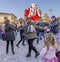 Blonde girl observes adults dancing in a circle with an allegorical float in the background during the parade of the Carnival