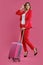 Blonde girl in red pantsuit, white blouse, high black heels. She smiling, posing with suitcase on pink background