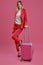 Blonde girl in red pantsuit, white blouse, high black heels. She smiling, posing with suitcase on pink background