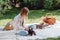 Blonde girl sitting on white blanket in garden and having picnic at sunny day with cute puppies.