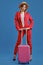 Blonde girl in straw hat, white blouse, red pantsuit, high black heels. She is leaning on a handle of pink suitcase