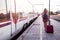 Blonde girl with summer clothes walks by the train with her red suitcase, before traveling