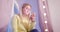 A blonde girl in a yellow sweater is sitting with a telephone in her room against a background of pink curtains and