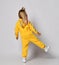 Blonde kid baby girlwith stylish hairstyle in modern yellow jumpsuit and sneakers stands back to camera on one leg