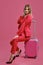Blonde lady in red pantsuit, white blouse, high black heels. She smiling, sitting on her suitcase on pink background
